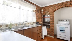 A kitchen with a brick wall, a built-in oven, a stove, and a window with lace curtains. - Villa Fiore- South Fremantle Retro Style (Beaconsfield)