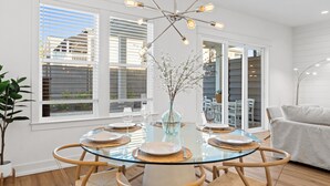 Un comedor con una mesa de vidrio rodeada de sillas de mimbre, una lámpara de araña moderna y vistas al exterior a través de amplios ventanales.