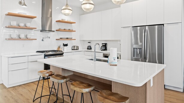 Une cuisine moderne avec des meubles blancs, des appareils en acier inoxydable, des tabourets de bar en bois et des luminaires suspendus.