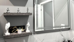 Une salle de bain moderne avec un lavabo en marbre, des toilettes et un grand miroir. Des objets décoratifs sont disposés sur des étagères murales au-dessus du miroir.