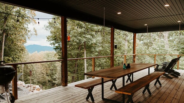 A wooden deck with a picnic table and chairs, overlooking a forested area with mountains in the distance.