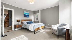 A bedroom with a bed, a chair, a nightstand, a lamp, a painting, and a window. - 1803 Historic Apartment in Heart of Salem•Steps to Haunted Happenings! (Salem)