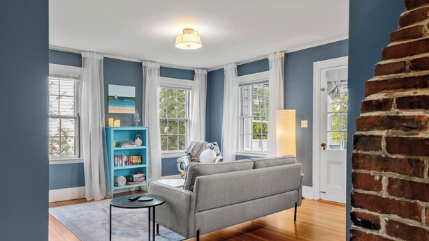 A room with a grey sofa, a small round table, a bookshelf, and a large window with curtains. - 1803 Historic Apartment in Heart of Salem•Steps to Haunted Happenings! (Salem)