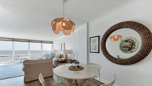 A modern living room with a round dining table, beige sofa, wicker chairs, and a large mirror on the wall.