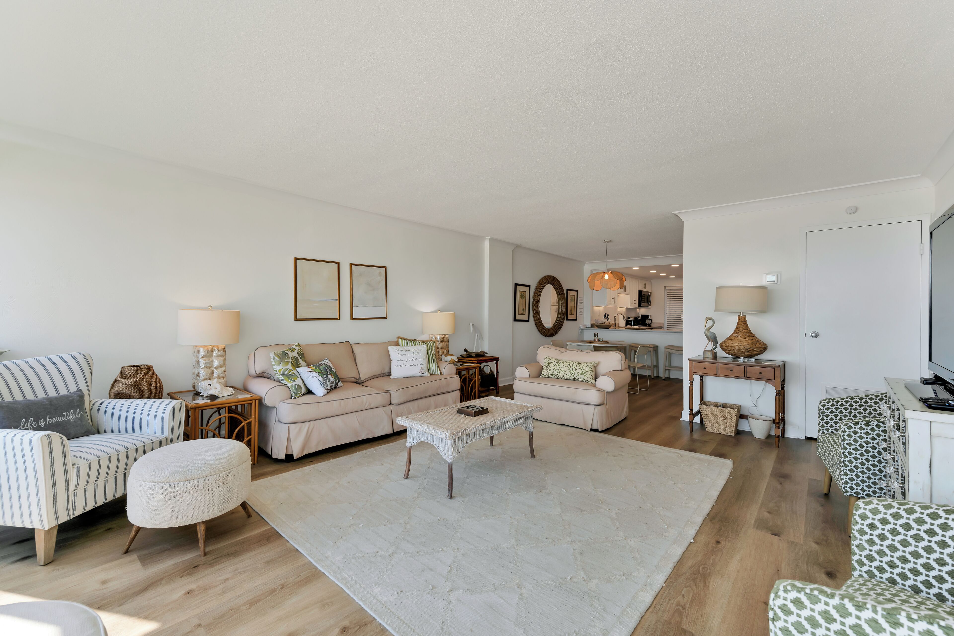 A spacious living room with a large sofa, two armchairs, a coffee table, and a TV. There are two lamps, a rug, and a dining area in the background.