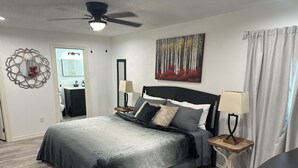 Un dormitorio ordenado con una cama, dos mesitas de noche, un ventilador de techo, un cuadro y un baño visible a través de un puerta abierta.