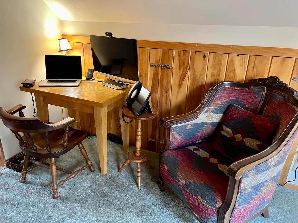 Ein Zimmer mit Schreibtisch aus Holz, Laptop, Stuhl und einem gemusterten Sessel.