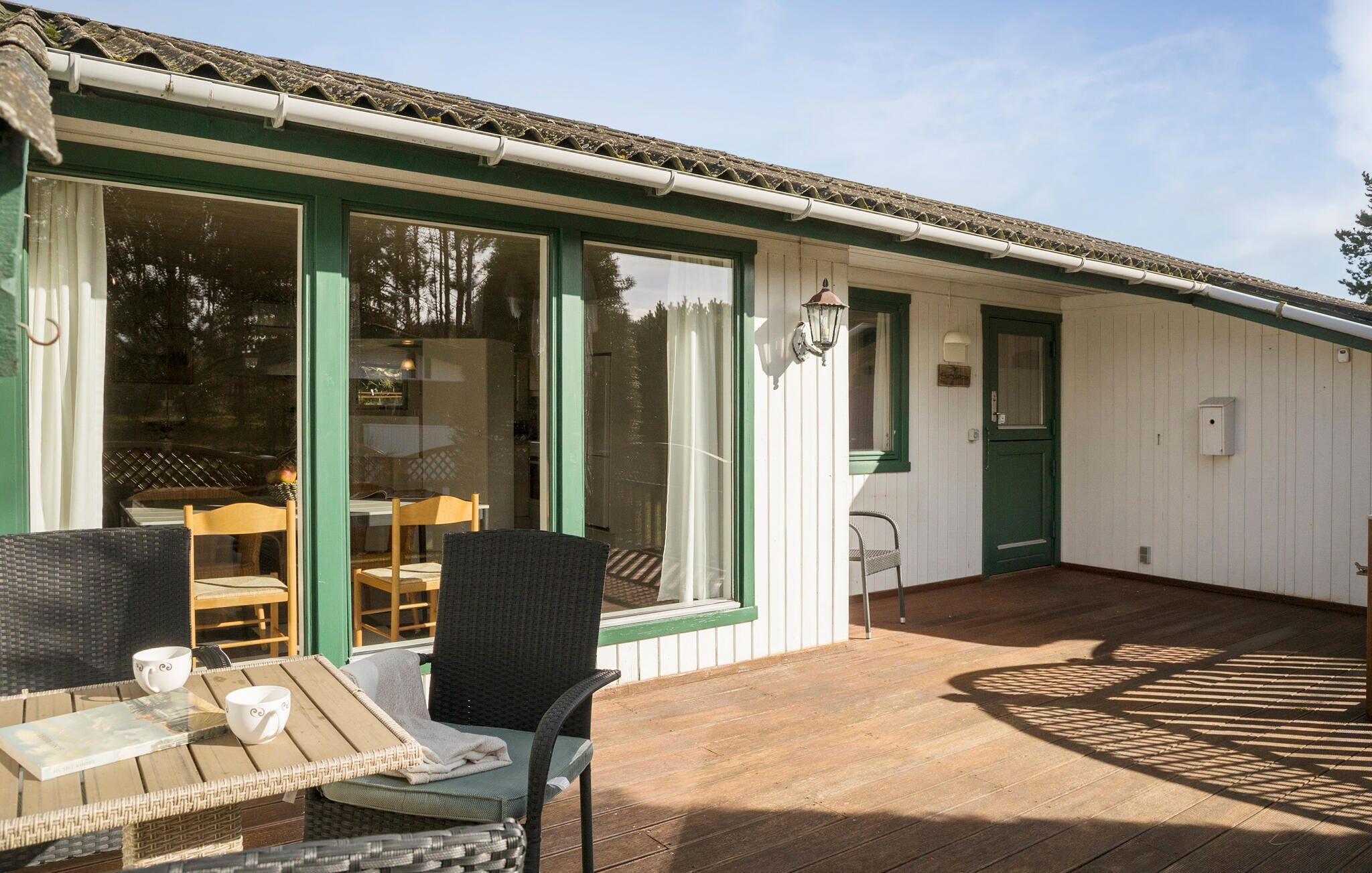 Een overdekt terras met rieten stoelen en een tafel, een groene deur en een raam met uitzicht op een terras.