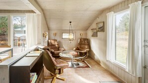 A cozy room with wooden walls and ceiling, featuring two leather chairs, a small round table, and a desk with a chair. There are windows with curtains, a chandelier, and framed pictures on the wall. - 3 bedroom stunning home in Rødby (Rødby)