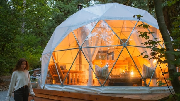 A geodesic dome structure with a wooden platform, interior lighting, and a view of the surrounding forest.