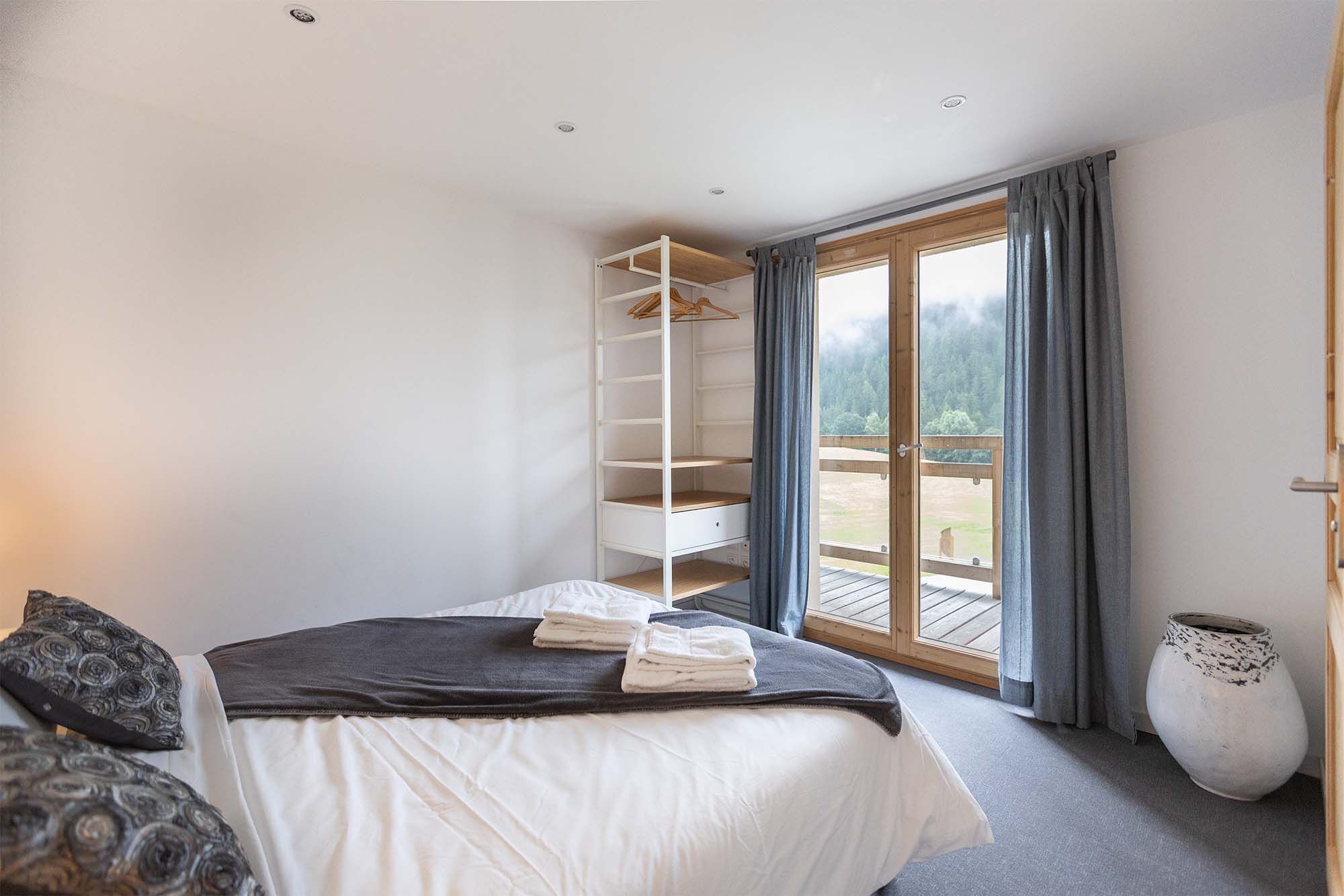A bedroom with a bed, a wardrobe, a large window with curtains, and a view of a forest.