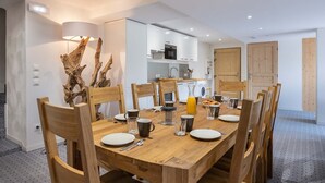 Un coin repas moderne avec une table en bois dressée pour quatre personnes, entourée de chaises en bois. Une grande plante décorative se trouve à gauche, et on aperçoit une cuisine au fond.