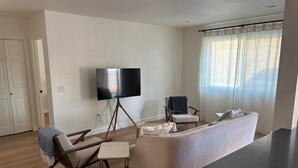A living room with a flat-screen TV on a stand, a sofa, a chair, a small table, and a window with curtains. - Modern Guesthouse on 5 Acre Estate Near Wineries (Temecula)