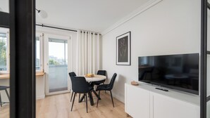 A modern living room with a large flat-screen TV, a dining area with a round table and black chairs, and a balcony with white curtains. - Aleja Waszyngtona Deluxe Apartment (Warszawa)