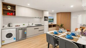 Een moderne keuken met witte kasten, een ingebouwde vaatwasser en een fornuis. Er is een eethoek met een tafel gedekt voor twee personen, met blauwe borden en glazen.