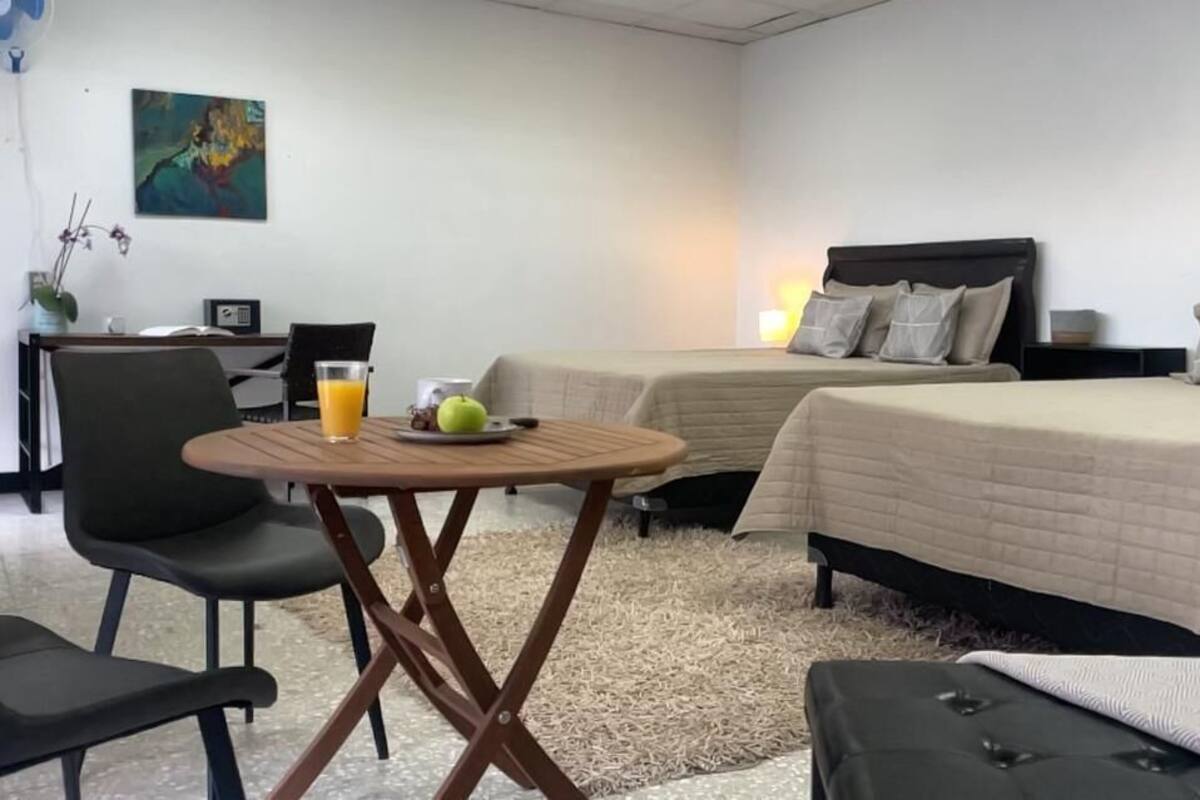 Una camera d'albergo con due letti, un tavolino con bevande e frutta, una sedia e una scrivania con una lampada.
