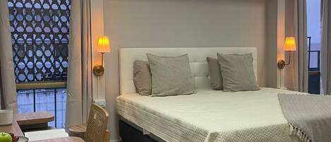 A hotel room with a bed, bedside lamps, a table with a chair, and a view through a window with a lattice screen.