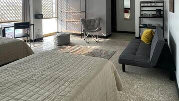 Un cuarto de hotel con una cama grande, un sofá gris con un cojín amarillo, un sillón pequeño, un escritorio y un televisor montado en la pared.