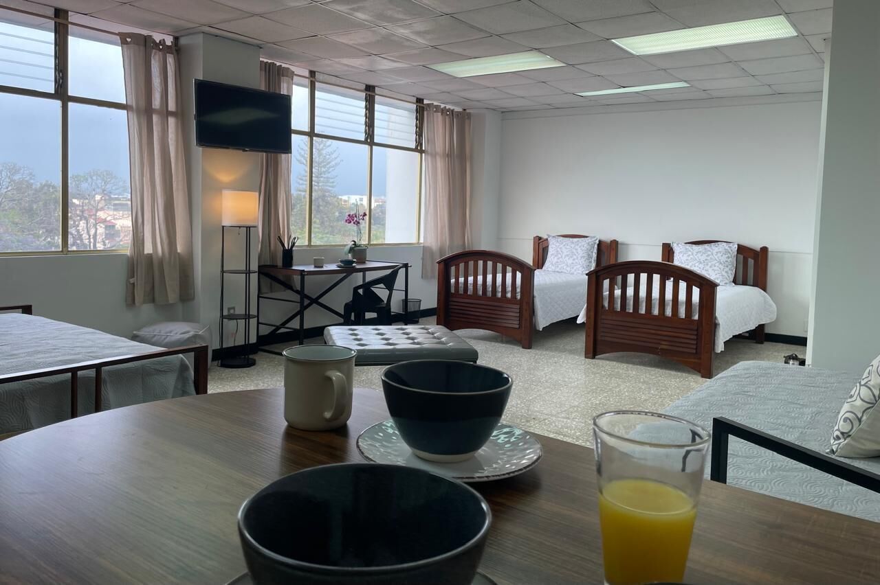 A hotel room with a bed, a desk with a chair, a table with bowls and a glass of orange juice, and a TV on the wall.