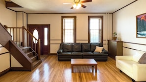 A living room with a wooden floor, a black leather sofa, a white armchair, a wooden coffee table, a ceiling fan, and a staircase. - Charming, historic 3-bedroom house in quaint Fredericksburg with AC, WiFi (Fredericksburg)