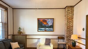 A cozy living room with a dark leather sofa, a white armchair, a wooden coffee table, a painting of a mountain landscape, and a brick accent wall. - Charming, historic 3-bedroom house in quaint Fredericksburg with AC, WiFi (Fredericksburg)