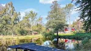 A wooden dock extending into a calm pond surrounded by trees and a small building with a red roof.