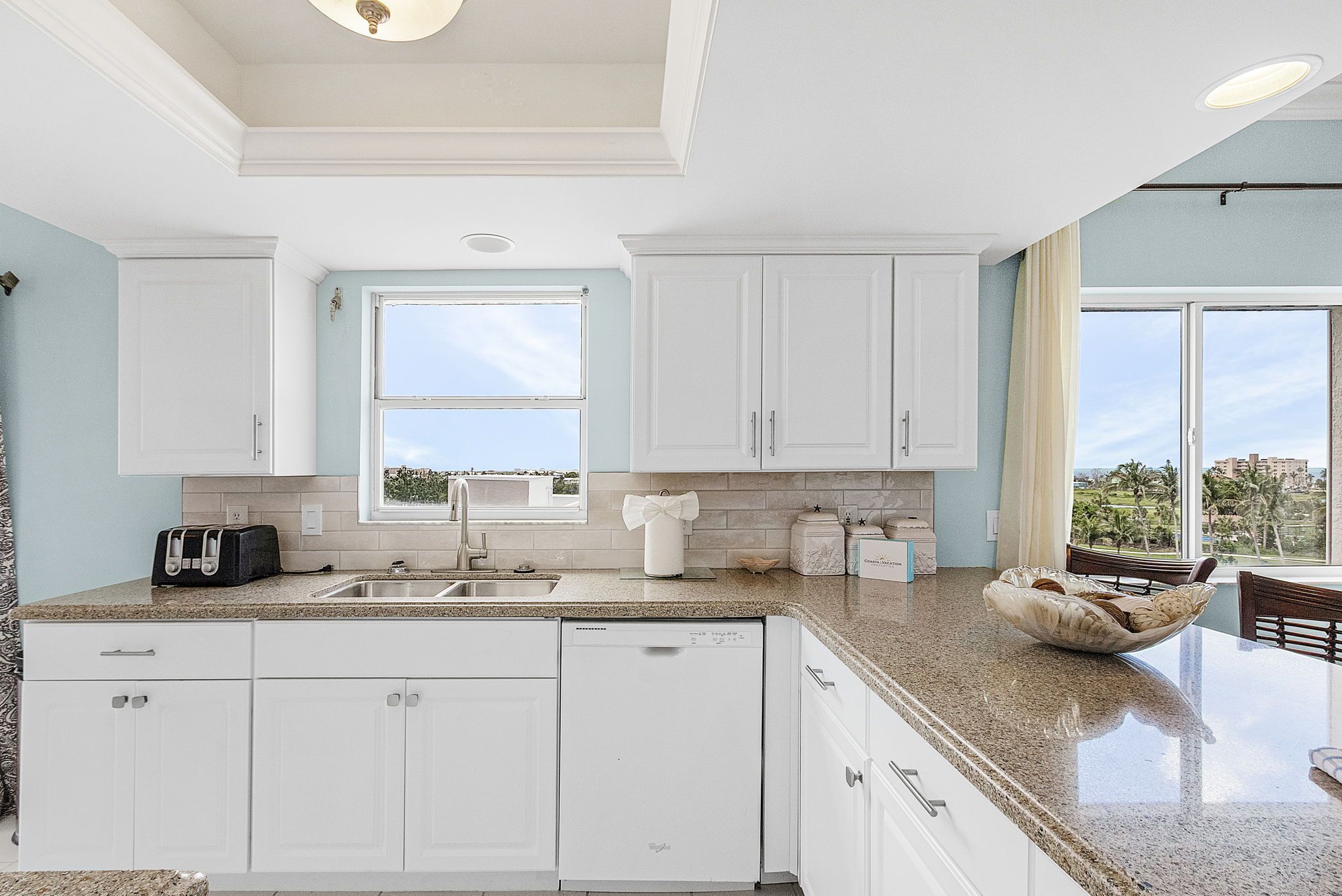 A modern kitchen with white cabinetry, granite countertops, and a large window offering a view of palm trees.