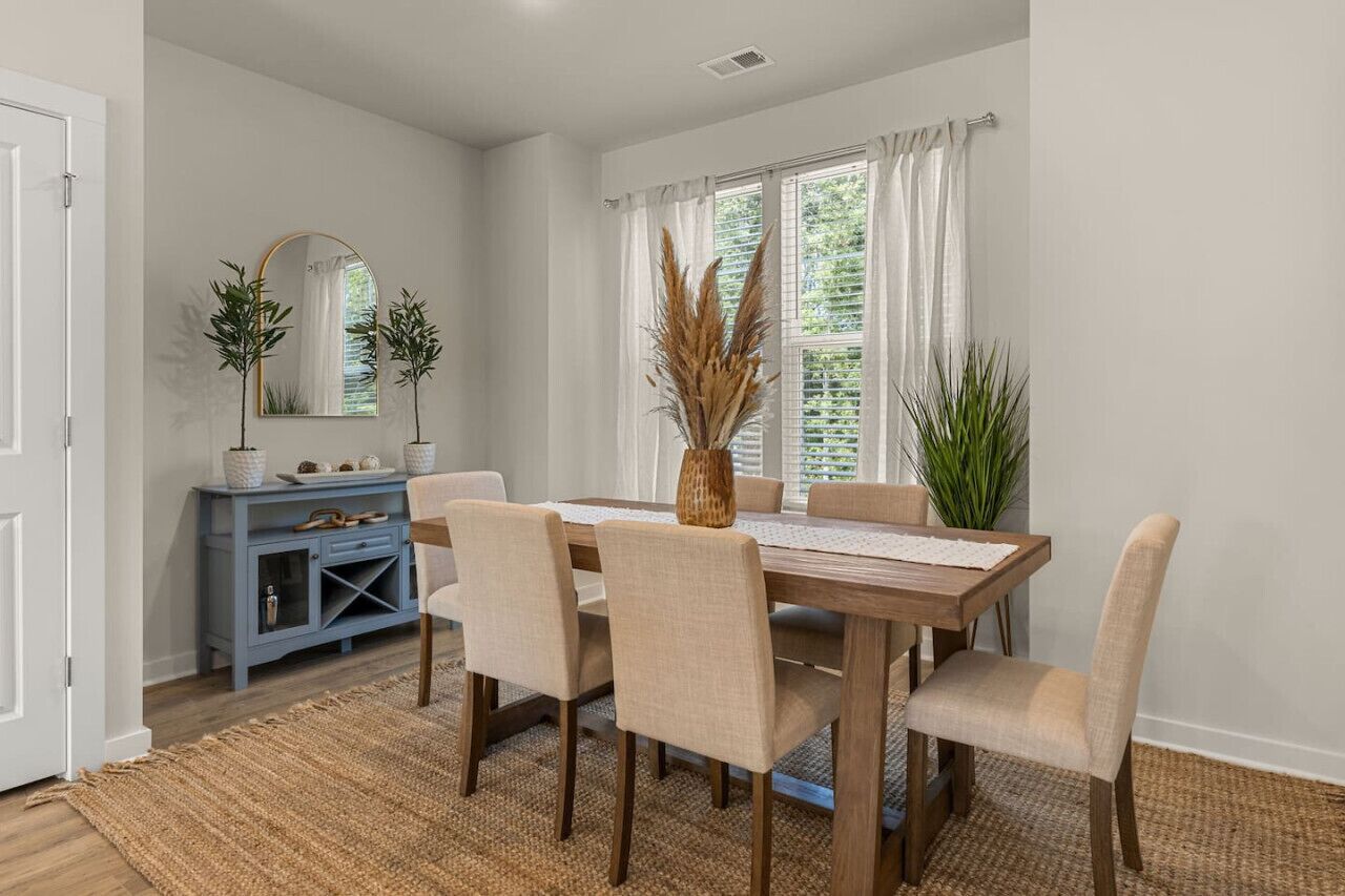 A dining area with a wooden table, six chairs, a blue console, and a large window with curtains.