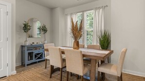 A dining area with a wooden table, six chairs, a blue console, and a large window with curtains. - Lux Charleston Residence for 10+ (Hanahan)