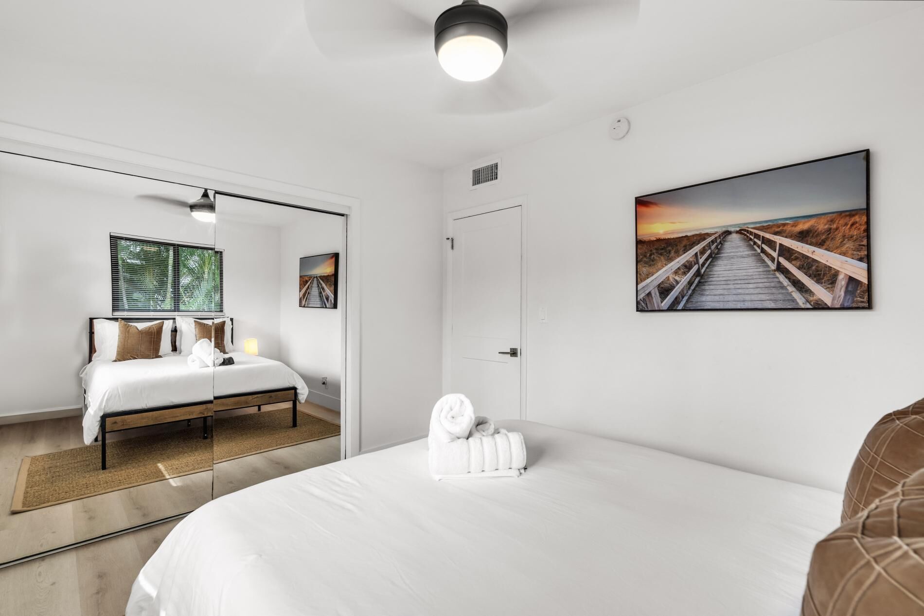 Ein modernes Hotelzimmer mit einem großen Bett, einem Nachttisch, einem Spiegel, einem Fenster mit Jalousien, einer Tür und einem Wandbild von einem Strandweg.