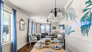 A dining area with a wooden table set for four, a ceiling fan, and a wall with a tropical leaf design and the word "sand to sea". - Beachfront | Direct Access, Outdoor Bar & Grill (Crystal Beach)
