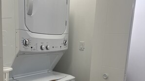 A compact laundry room with a front-loading washer and dryer, a small trash bin, and a door leading to another room. - Modern & Central 3BED/2BATH near Health Science Center (Winnipeg)