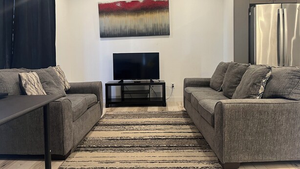 A modern living room with two grey sofas, a flat-screen TV on a black stand, a large abstract painting on the wall, and a kitchen area with a stainless steel refrigerator. - Modern & Central 3BED/2BATH near Health Science Center (Winnipeg)
