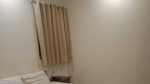 A single bed with white bedding, a nightstand with a lamp, and a window with beige curtains. - Gatsby Hotel Jaipur (Jaipur)