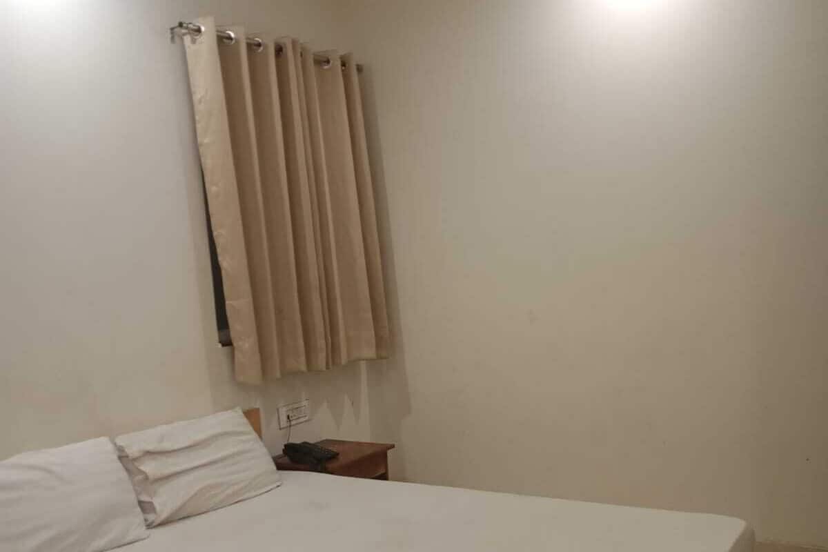 Una cama individual con ropa blanca, una mesita de noche con lámpara y una ventana con cortinas color beige.