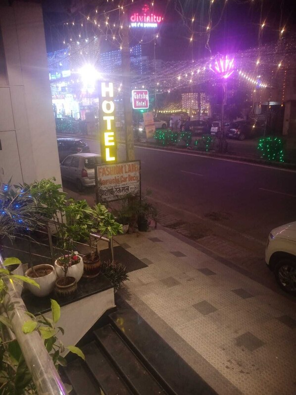 Entrada de hotel con un letrero de neón, un anuncio para un laboratorio de moda y plantas en macetas.