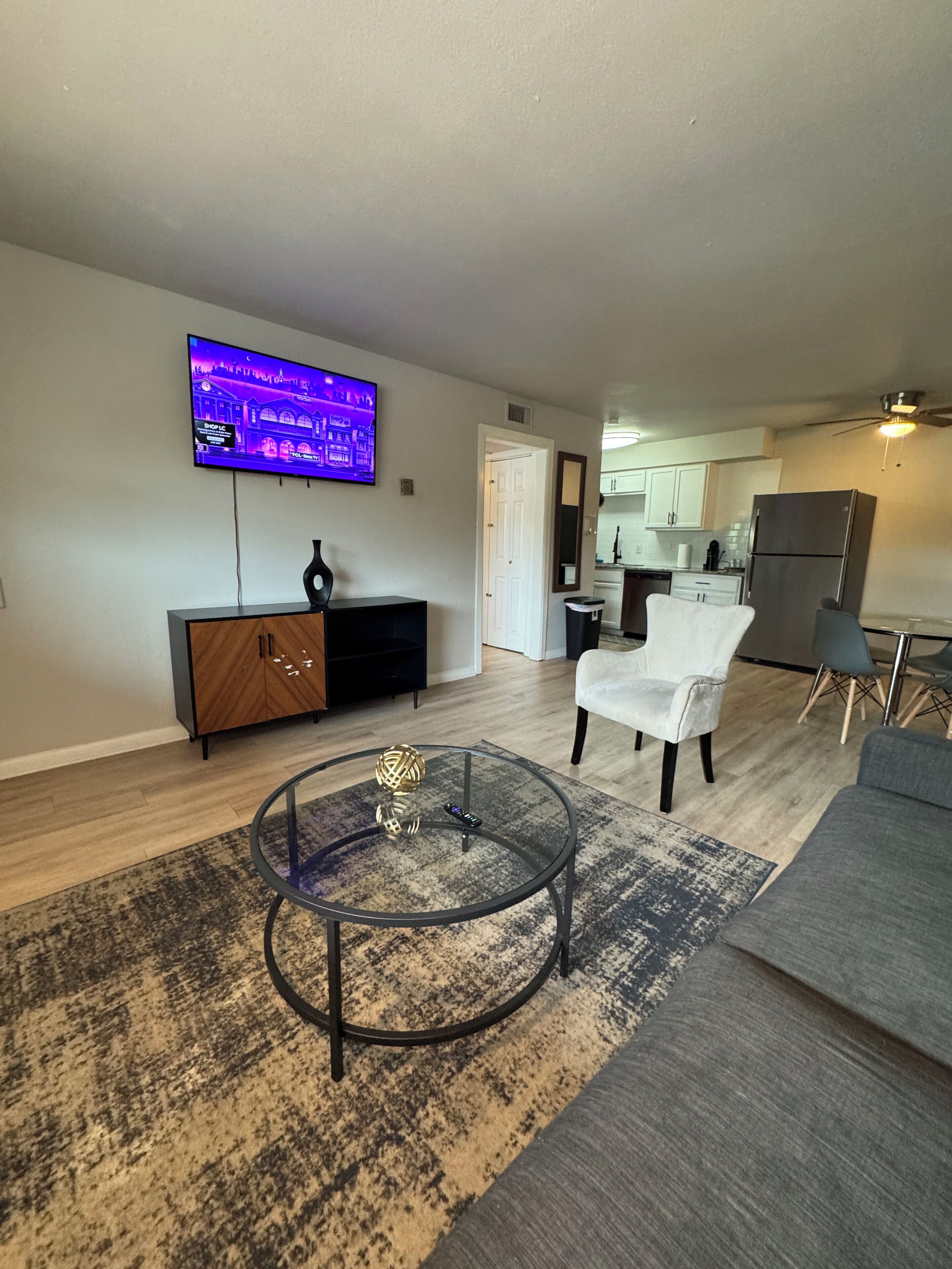 A modern living room with a flat-screen TV mounted on the wall, a glass coffee table, a grey sofa, and a kitchen in the background.