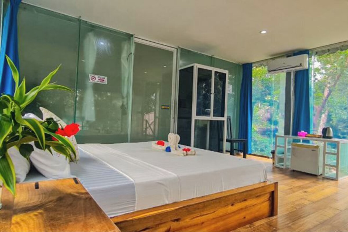 A modern hotel room with a large bed, a wooden headboard, a small desk, and a large window offering a view of greenery.
