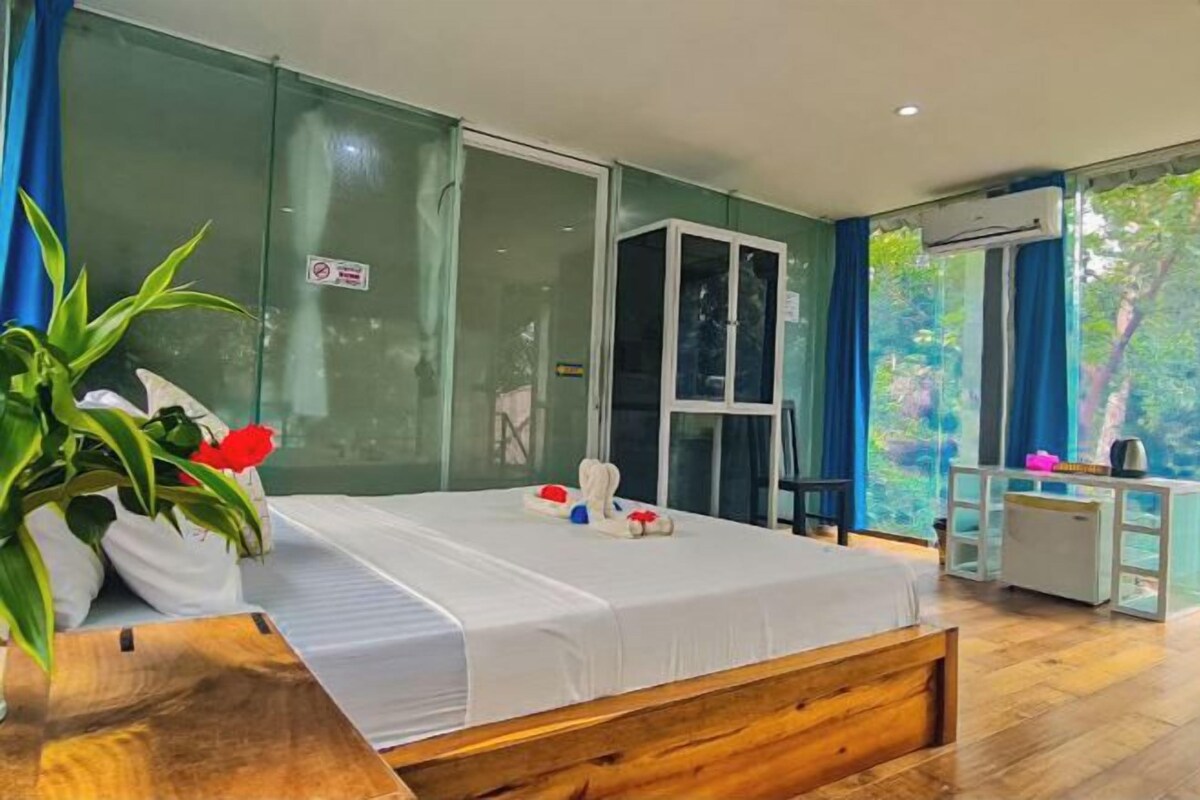 Une chambre d’hôtel moderne avec un grand lit, une tête de lit en bois, un petit bureau et une grande fenêtre donnant sur un paysage verdoyant.