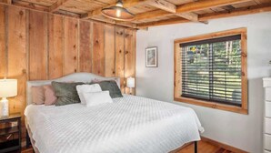 A bedroom with a wooden ceiling, a bed with pillows, a nightstand, a window with blinds, and a picture on the wall. - Relax. Soak. Explore. Repeat. | Bella Luna Cabin (Ruidoso)