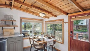 A dining area with a wooden table and chairs, a kitchen with grey cabinets, and large windows with a view of greenery. - Relax. Soak. Explore. Repeat. | Bella Luna Cabin (Ruidoso)