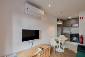 Une cuisine moderne dotée d’un four encastré, un coin repas avec une table ronde et des chaises, un téléviseur à écran plat fixé au mur et un banc en bois avec une télécommande.