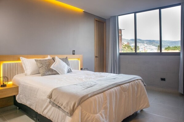A modern bedroom with a large bed, a nightstand, a window with a view of a cityscape, and a wooden headboard.