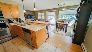 A modern kitchen with wooden cabinets, a stainless steel stove, and a dishwasher. There is a dining area with a table and chairs, and a large window providing a view of the outdoors. - 2-bedroom condo with hot tub and mountain views (Golden)