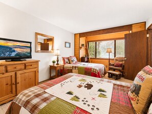 A cozy bedroom with a television, a bed with a quilt, a chair, and a window with blinds. - Mama Bear's Cottage - Portable AC, Close to Slopes, Fenced Yard, Pet Friendly (Big Bear Lake)
