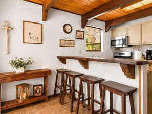 Een keuken met houten barstoelen, een klok, een bord met opschrift 'koffie' en een raam met jaloezieën.