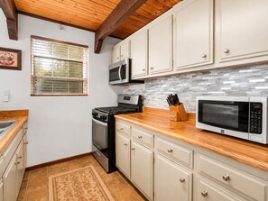 A kitchen with white cabinets, a microwave, a stove, and a window with blinds. - Mama Bear's Cottage - Portable AC, Close to Slopes, Fenced Yard, Pet Friendly (Big Bear Lake)