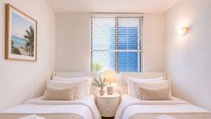 Une chambre d’hôtel avec deux lits, une table de chevet avec une lampe, une fenêtre avec des stores et un tableau encadré au mur.