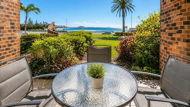 Une terrasse avec une table en verre, des chaises et un pot de fleurs, donnant sur un espace aménagé avec vue sur l’océan et les palmiers.