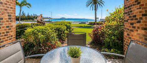 A patio with a glass table, chairs, and a potted plant, overlooking a landscaped area with a view of the ocean and palm trees.